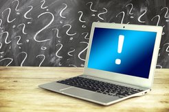 Laptop displaying an exclamation mark on its screen standing on a table in front of a blackboard covered in question marks